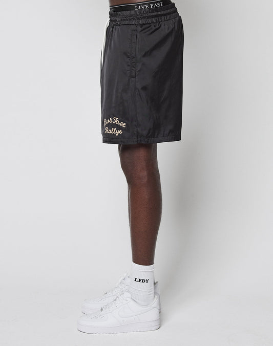 A person in profile wears black LFDY Retro Satin Shorts by LIVE FAST DIE YOUNG, featuring a Chainstitch Logo and Live Fast Rallye text in an Oversize Fit, plus white LEDY socks and white Nike sneakers, against a plain white background.