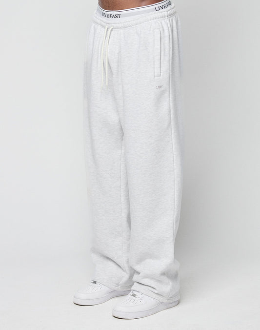 A person wearing Basic Wide Jogger sweatpants by LIVE FAST DIE YOUNG in light gray with an elastic waistband, drawstring, and bold logo print, paired with white sneakers, stands against a plain white background. | model