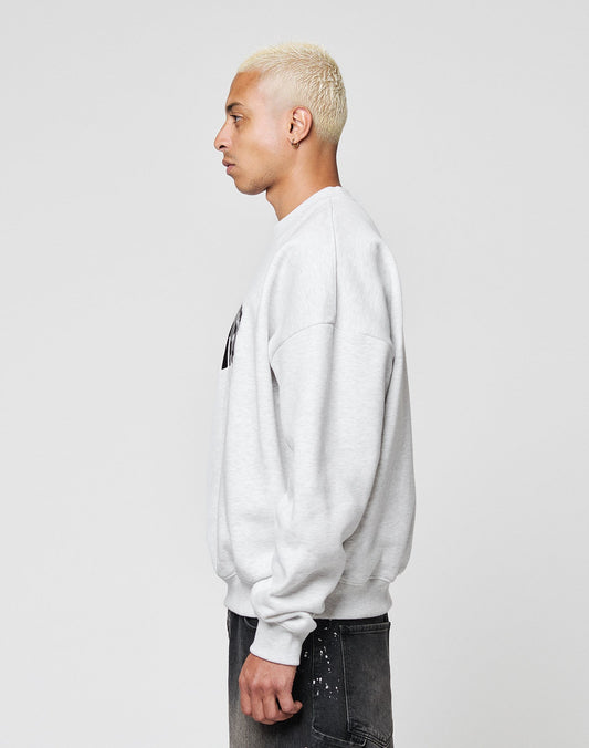 A person with short, bleached blond hair wears a light gray LFDY Emblem Sweater by LIVE FAST DIE YOUNG and dark jeans, standing in profile against a plain, light background.