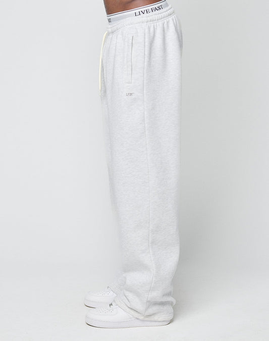 A person in profile wears LIVE FAST DIE YOUNG Basic Wide Jogger sweatpants in light gray, featuring a relaxed fit, subtle upper thigh logo, and elastic waistband, styled with white sneakers against a plain white background.