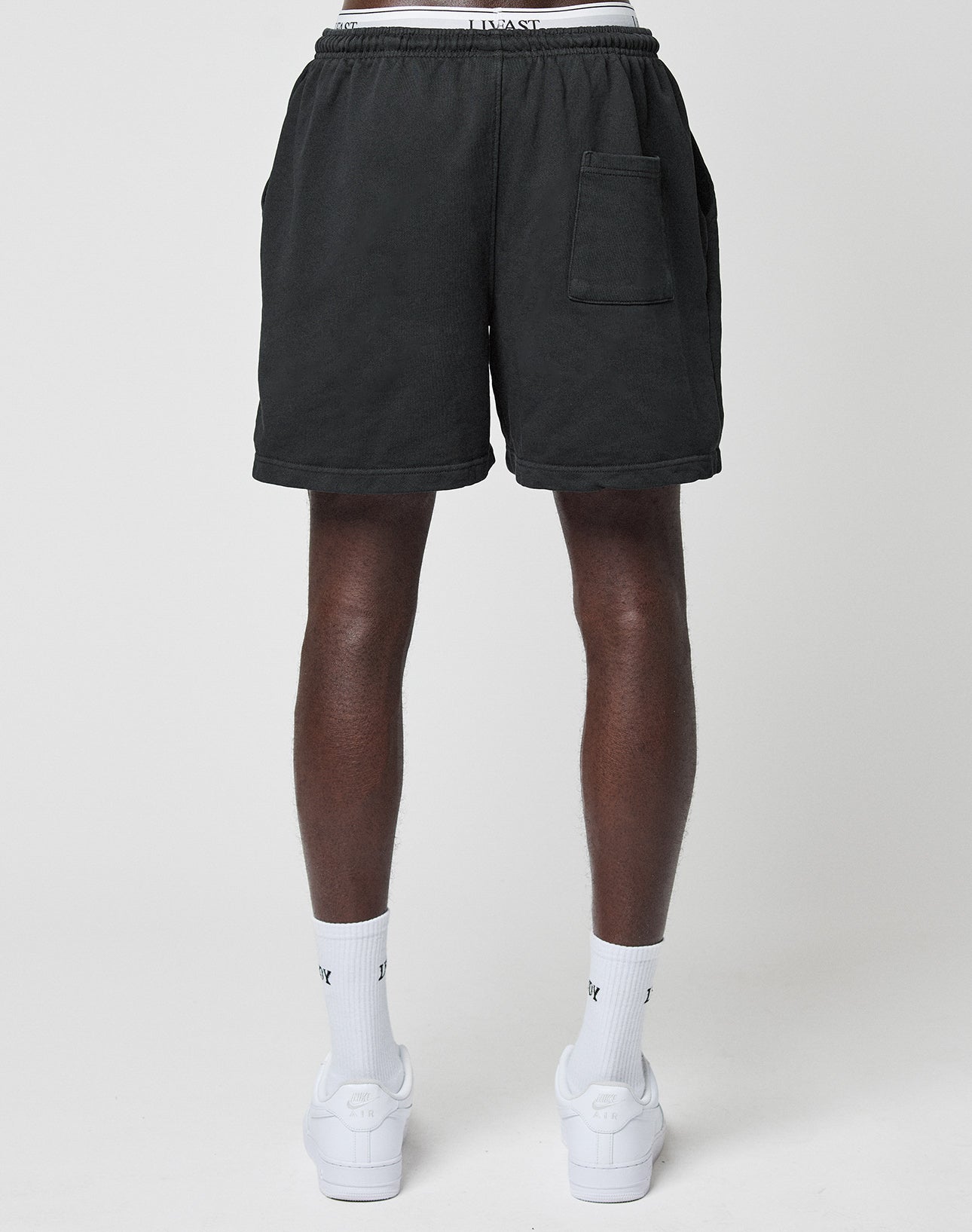 A person stands facing away, wearing LIVE FAST DIE YOUNG Basic Sweat Shorts in dark gray with an elastic waistband and back pocket, paired with white socks and sneakers against a plain light background.
