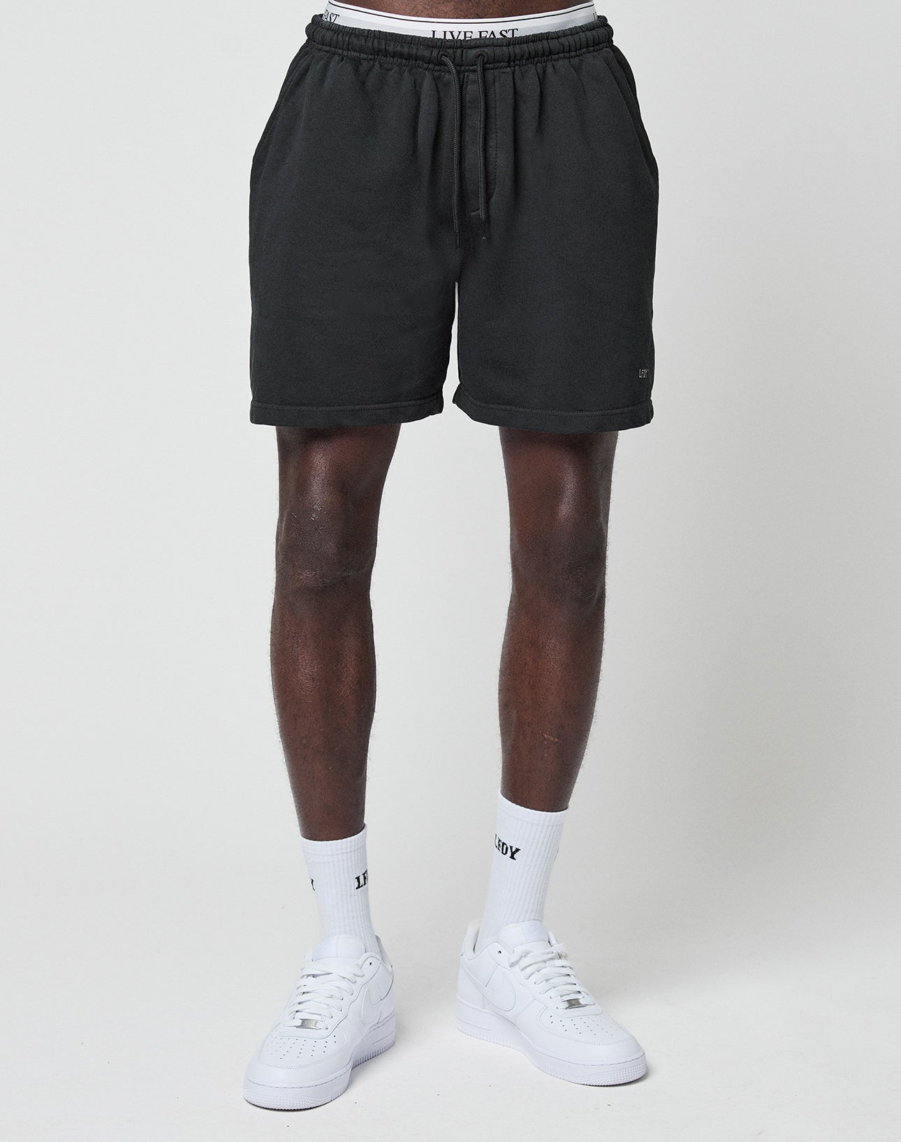 A person wears LIVE FAST DIE YOUNG Basic Sweat Shorts with an elastic waistband, white socks, and white sneakers, standing against a plain white background. The image is cropped from the waist to below the knees.