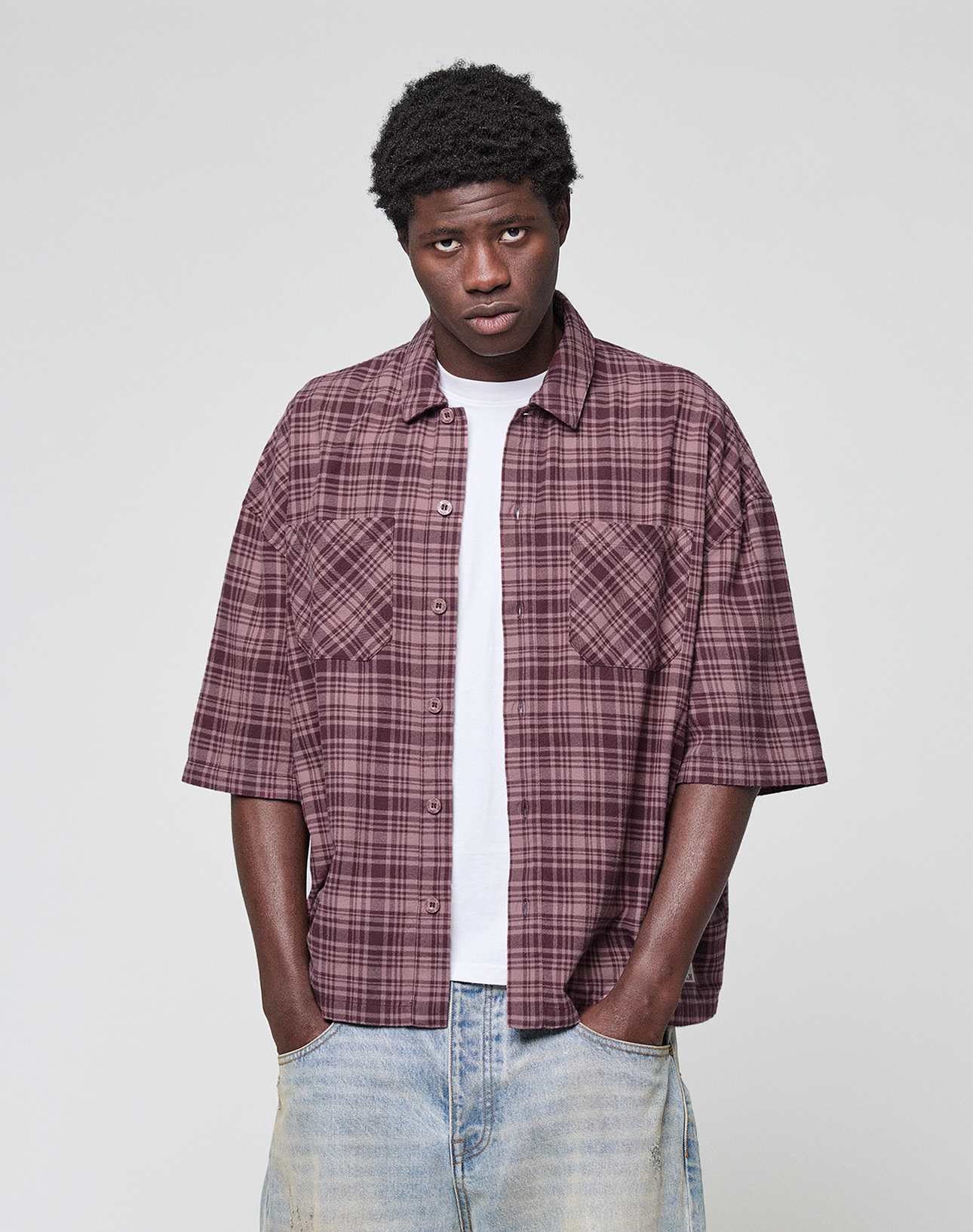 A man wears the LIVE FAST DIE YOUNG LFDY Light Flannel in maroon plaid over a white t-shirt and light blue jeans, standing against a light gray background with hands in pockets. Subtle LFDY-Branding completes the look.