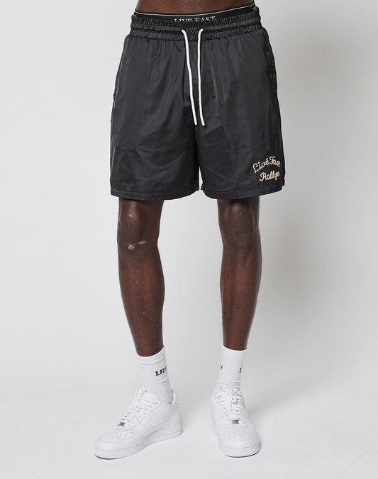 A person wears black LFDY Retro Satin Shorts by LIVE FAST DIE YOUNG, featuring a yellow chainstitch logo, along with white socks and sneakers, standing against a plain white background.
