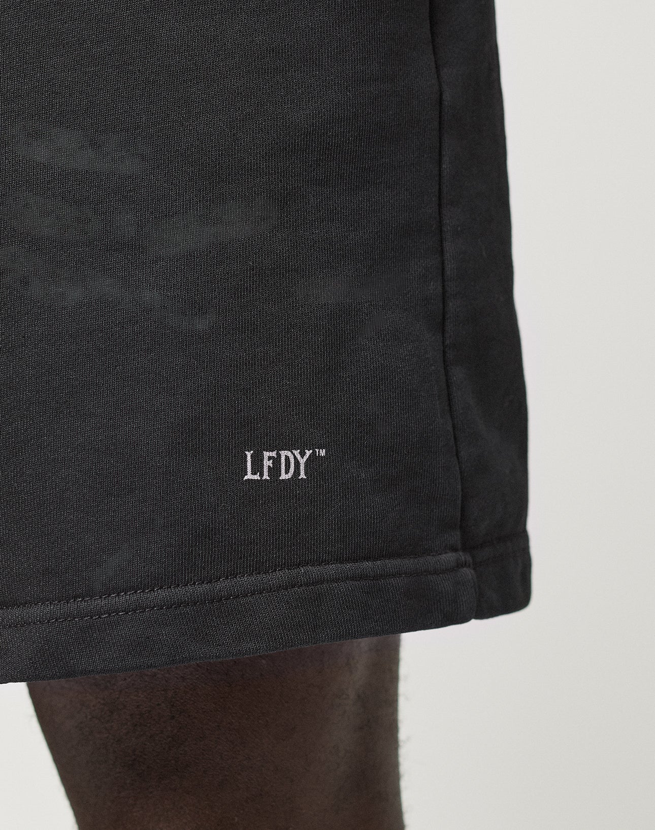 A close-up shows a person wearing the LIVE FAST DIE YOUNG Basic Sweat Shorts in black with an oversized fit and a white LFDY™ logo embroidered near the hem, set against a plain, minimal background.