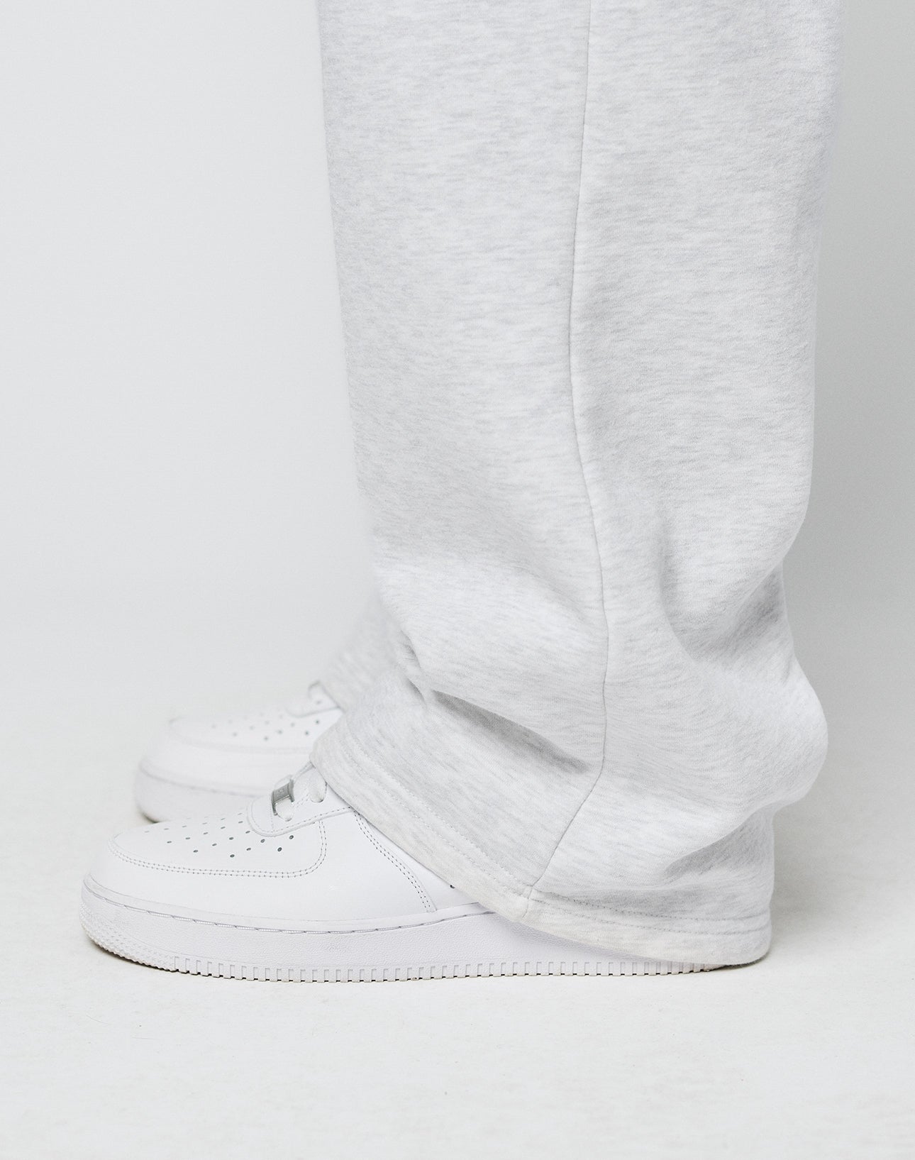 Close-up side view of a person wearing LIVE FAST DIE YOUNG Basic Wide Jogger in light gray with elastic waistband and drawstring, paired with white sneakers. The oversized fit slightly covers the shoes while standing on a white surface.