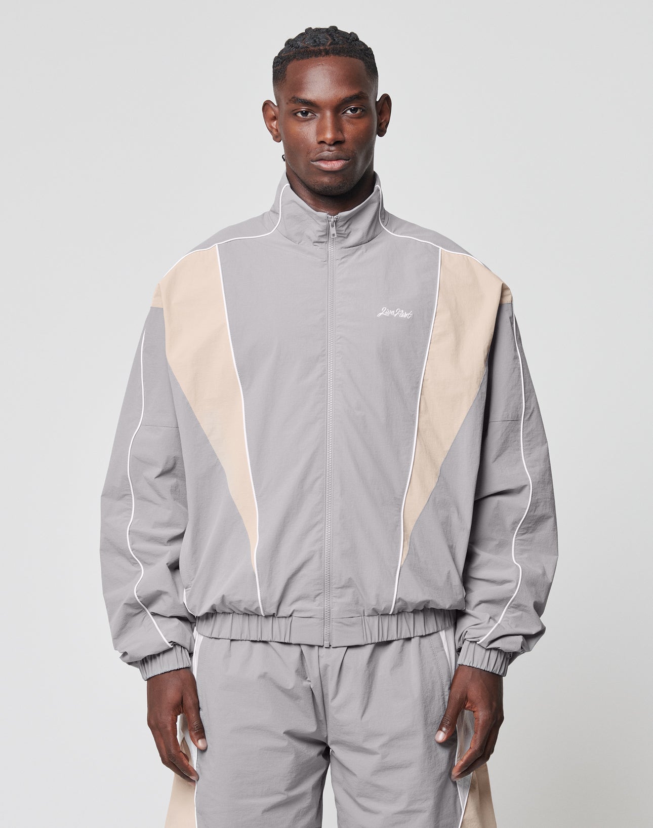 A man stands facing forward in a LIVE FAST DIE YOUNG Basic Nylon Trackjacket, regular fit, light gray and beige with white piping, made from nylon-lycra fabric, against a plain light gray background.