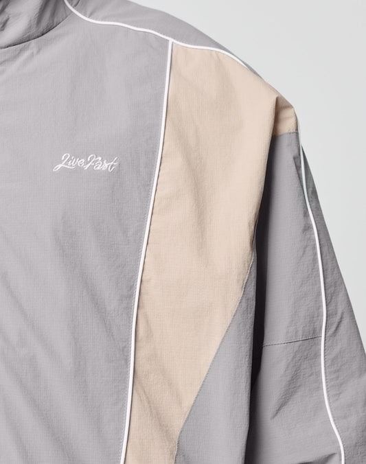 A close-up of the Basic Nylon Trackjacket by LIVE FAST DIE YOUNG, featuring light gray and beige color-blocking, white piping, Logo-Stick detail, and Live Fast embroidered in white on the chest.
