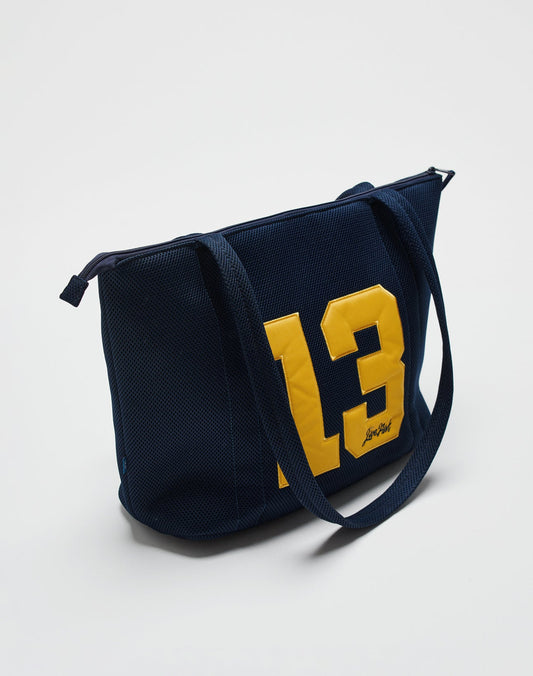 The LFDY Mesh Beach Bag by LIVE FAST DIE YOUNG is a navy blue tote with long handles, a secure zipper, a large yellow 13 and small signature on one side, shown against a plain white background.