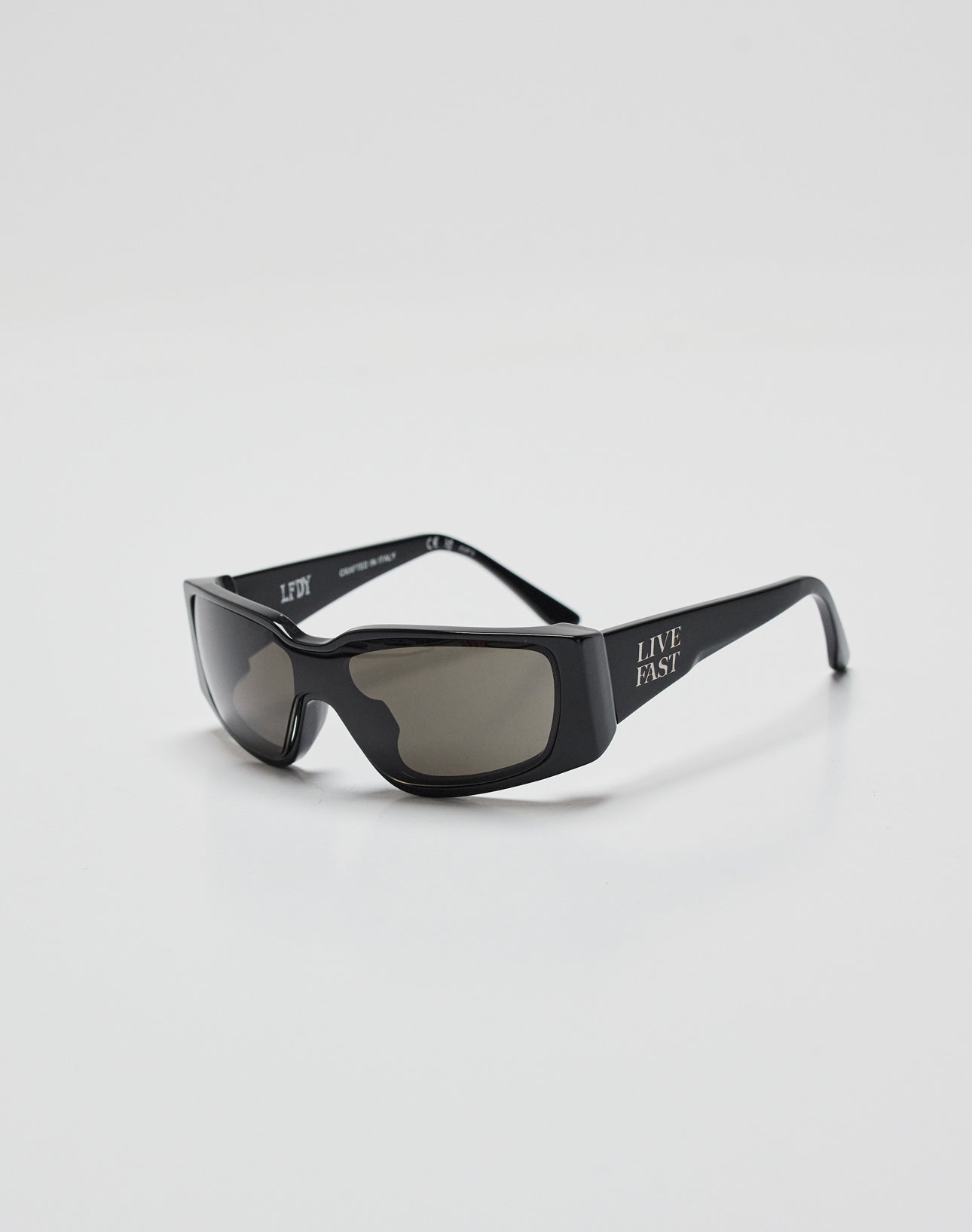 LIVE FAST DIE YOUNGs Live Fast Curved Shades are black rectangular sunglasses with dark lenses, made from Mazzuchelli Acetate, offering 400 UV protection and LIVE FAST in white on the left temple, shown on a plain white background.