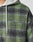 Close-up of the LIVE FAST DIE YOUNG Vintage Padded Hooded Flannel in green and black plaid with a light gray hood, drawstring, and visible Flag-Label tag, showing part of a person’s shoulder and chest.