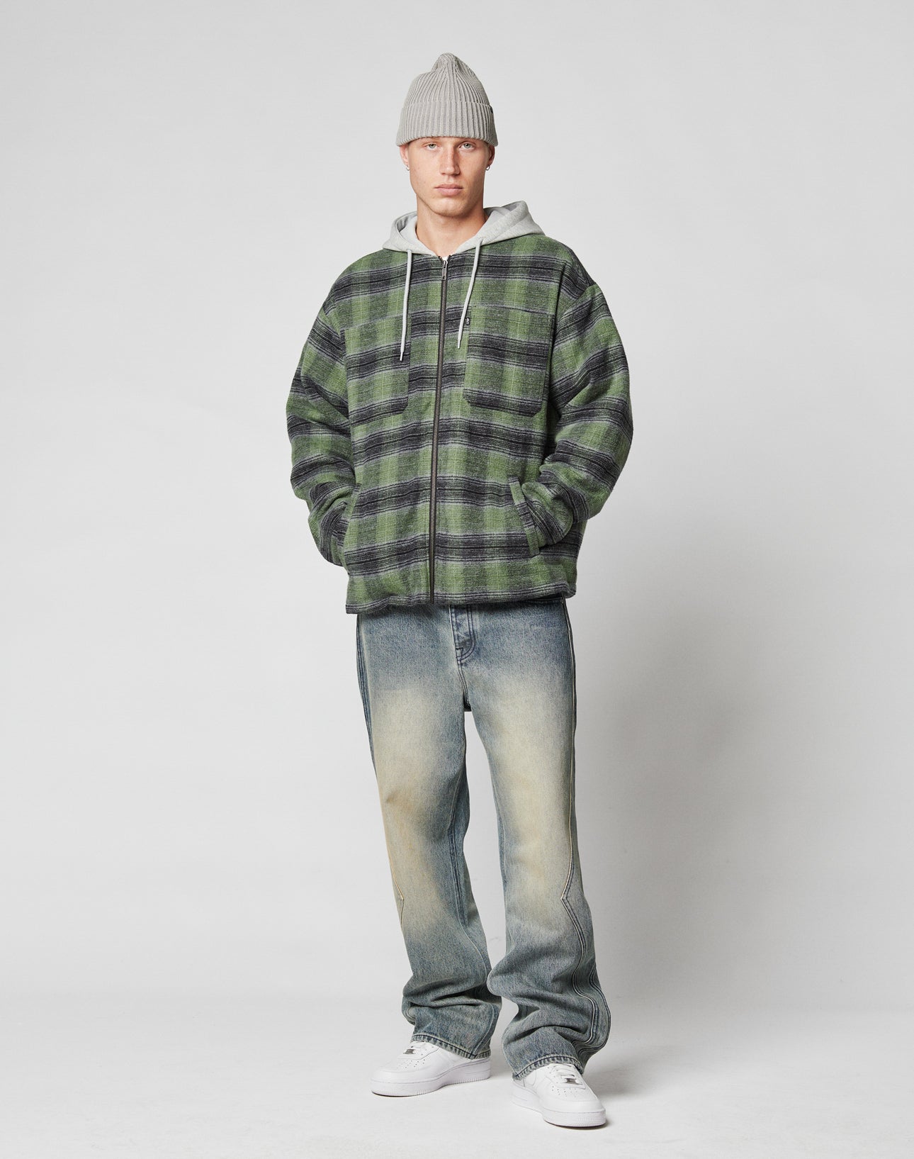 A person stands against a plain background wearing the LIVE FAST DIE YOUNG Vintage Padded Hooded Flannel, a gray beanie, faded blue jeans in vintage wash, and white sneakers with hands in the flannel’s hoodie pockets.