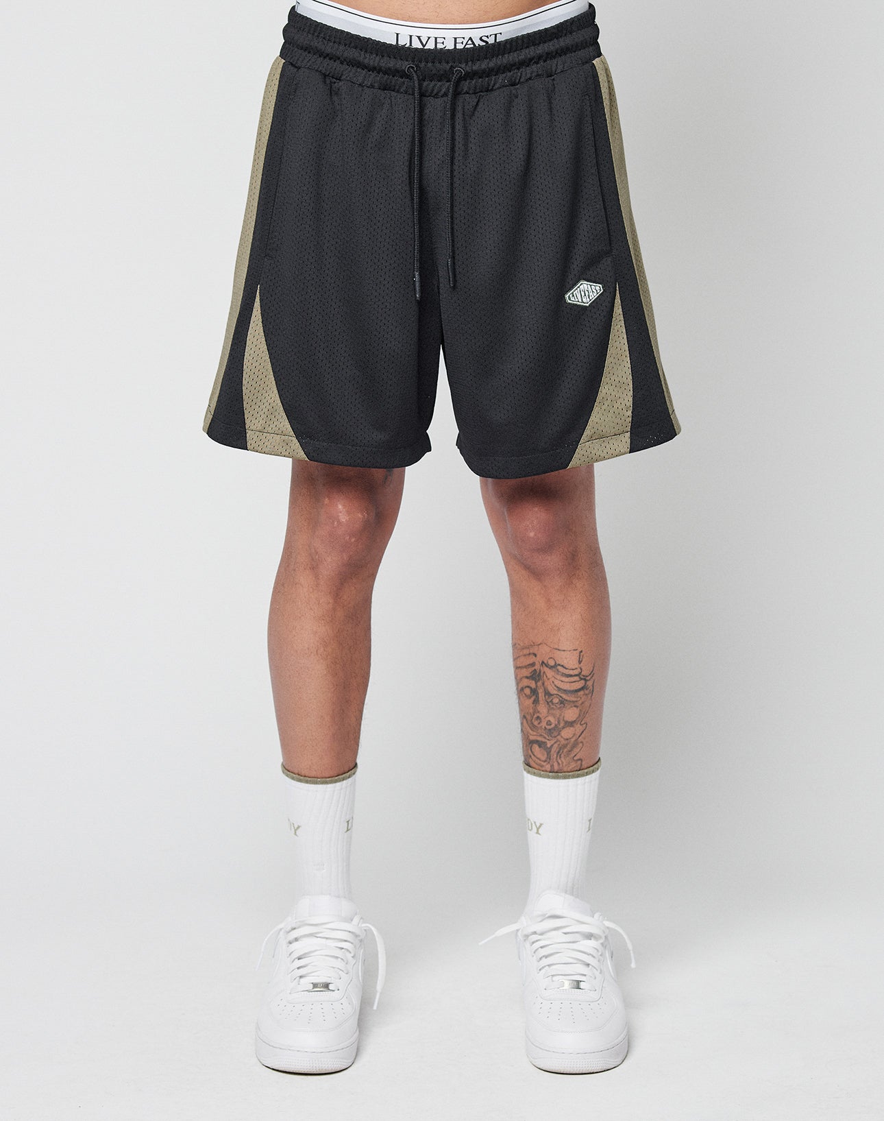 Someone stands against a plain background, showing only their lower torso and legs, wearing LIVE FAST DIE YOUNG Striped Mesh Shorts in black with olive side panels, a subtle logo patch, white socks, and white sneakers.