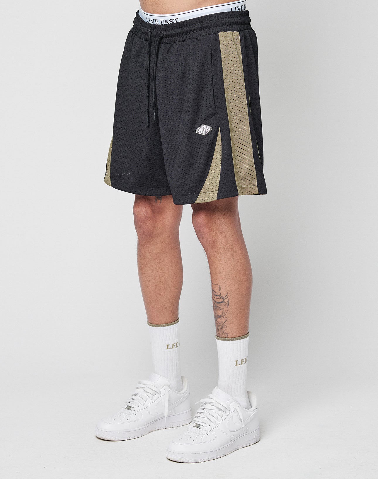 A person stands against a white background wearing LIVE FAST DIE YOUNG Striped Mesh Shorts—black with gold side stripes, white logo patch—paired with LIVE FAST text crew socks and white sneakers. The shorts have a regular fit and comfortable polyester feel. | model