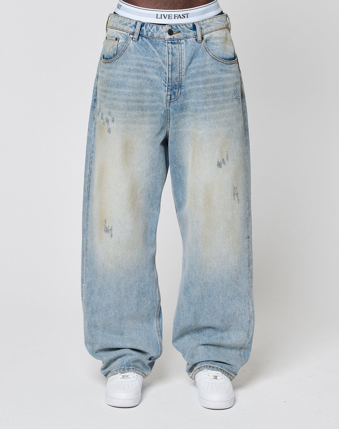 A person wears LIVE FAST DIE YOUNG’s LFDY Baggy Dirt Denim—baggy, light-wash blue jeans with faded and distressed details—paired with white sneakers and a branded waistband. The background is plain and light-colored.