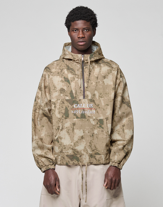 A man with short curly hair wears a LIVE FAST DIE YOUNG LFDY Camo Halfzip Jacket in oversize fit with "CALL US 449Livefast" on the front, paired with light beige 100% Baumwolle pants, standing against a light gray backdrop. | model