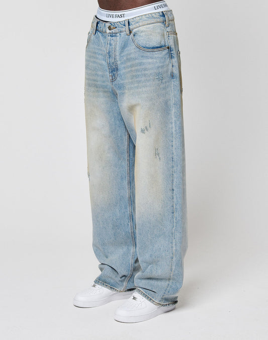 A person wears light blue LFDY Baggy Dirt Denim jeans by LIVE FAST DIE YOUNG, featuring a relaxed, baggy fit, subtle front fading, gerades Bein, and a visible white LIVE FAST waistband, paired with white sneakers. Background is plain white.