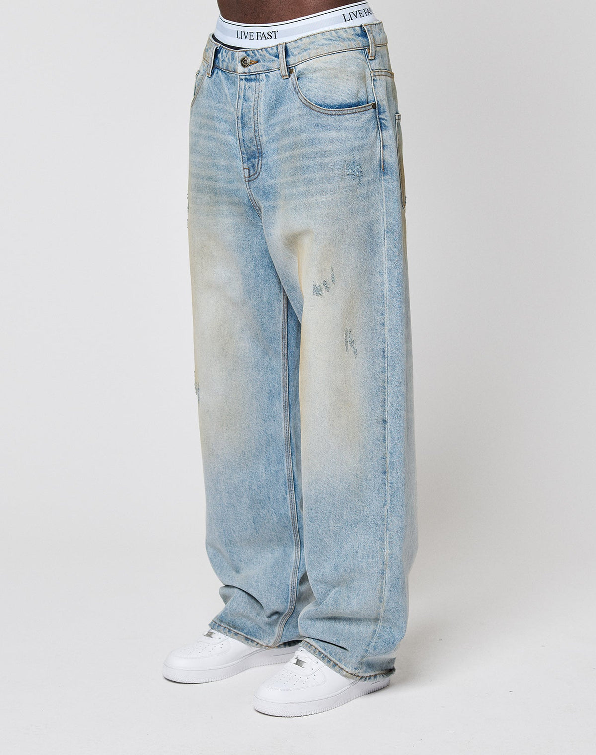 A person wears LFDY Baggy Denim by LIVE FAST DIE YOUNG—light-wash, subtly distressed jeans—with white sneakers and a white waistband featuring LIVE FAST branding, standing against a plain white background.| model