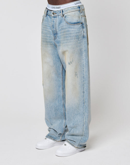 A person wears LFDY Baggy Denim by LIVE FAST DIE YOUNG—light-wash, subtly distressed jeans—with white sneakers and a white waistband featuring LIVE FAST branding, standing against a plain white background.| model