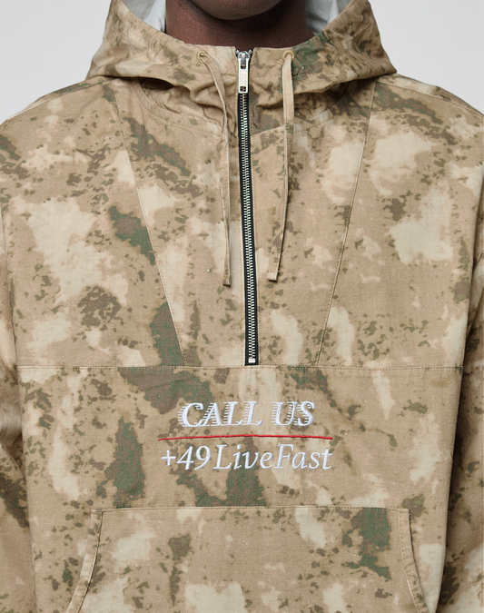 A person wears the LFDY Camo Halfzip Jacket by LIVE FAST DIE YOUNG—an oversized tan and green camo hoodie featuring a half-zip, embroidered "CALL US" (crossed out) and "+49 LiveFast" in white, gray, and red. Made from 100% Baumwolle.