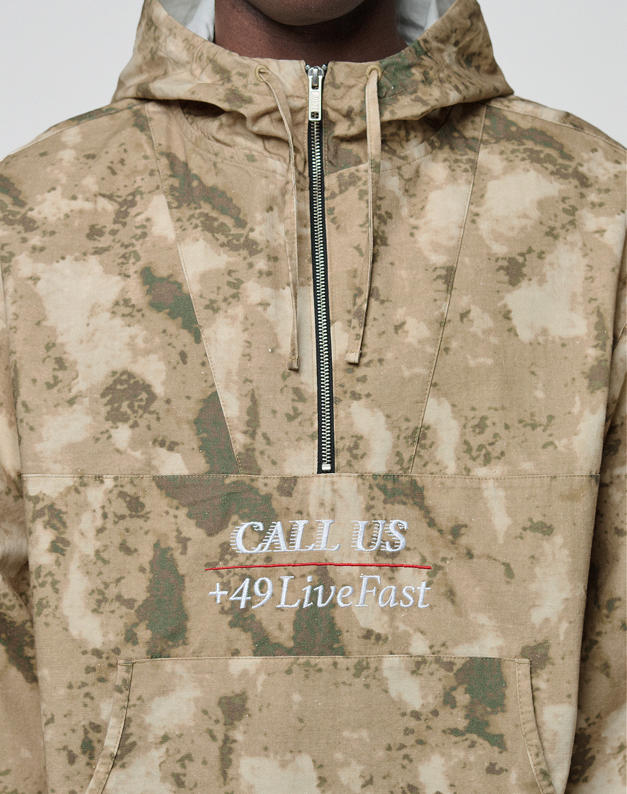A person wears the LFDY Camo Halfzip Jacket by LIVE FAST DIE YOUNG—an oversized tan and green camo hoodie featuring a half-zip, embroidered "CALL US" (crossed out) and "+49 LiveFast" in white, gray, and red. Made from 100% Baumwolle.