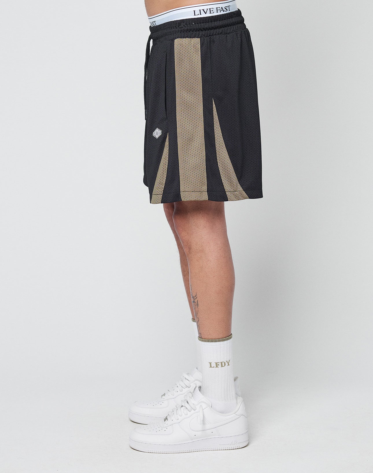 A person stands sideways in LIVE FAST DIE YOUNG Striped Mesh Shorts, showing a white waistband with LIVE FAST text, paired with white crew socks featuring LEDY text and white Nike sneakers. The background is plain white.
