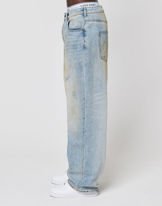 A person stands sideways in LFDY Baggy Denim by LIVE FAST DIE YOUNG—light-wash blue jeans with subtle distressing and a LIVE FAST waistband—paired with white sneakers against a plain white background.