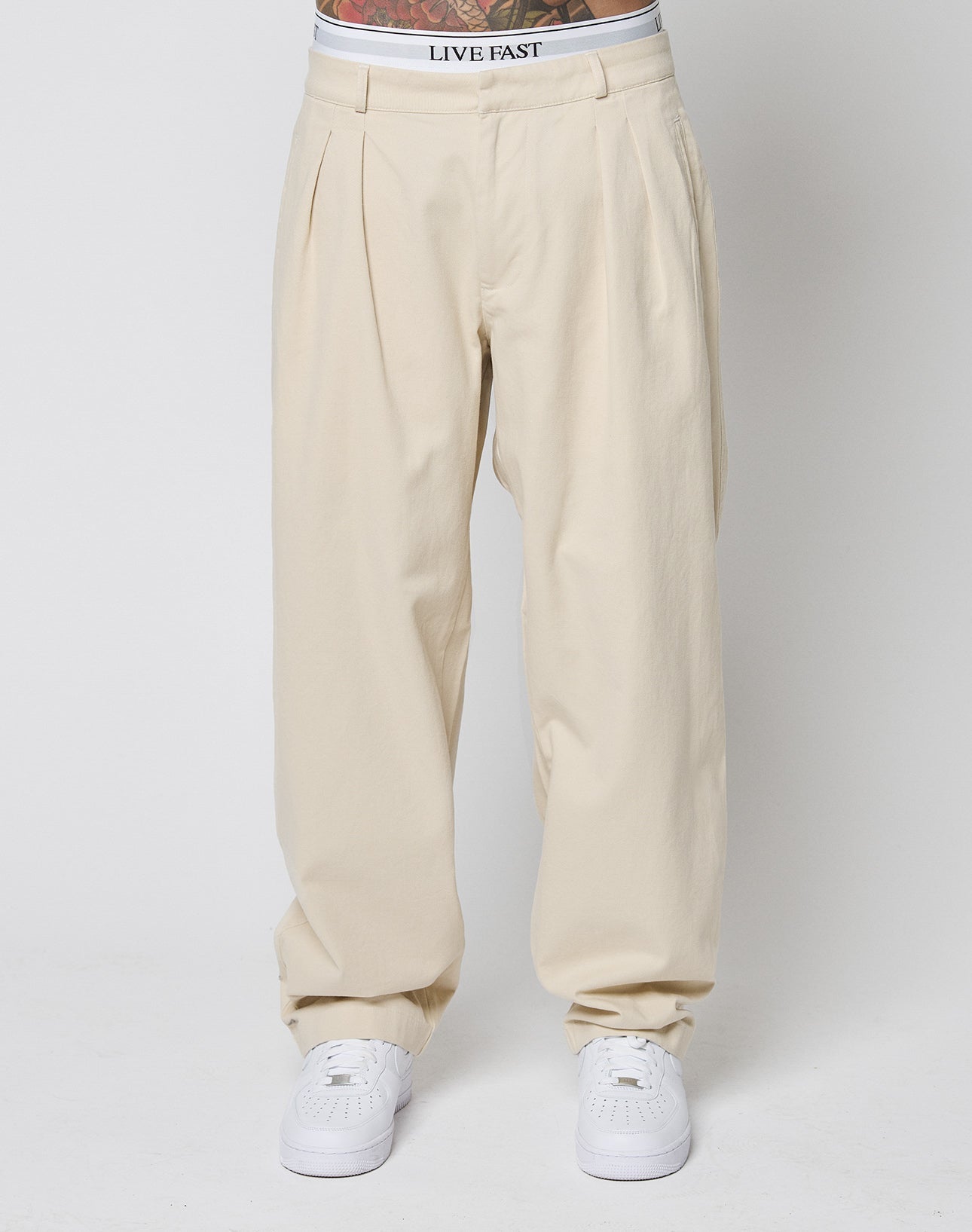 A person wears LIVE FAST DIE YOUNG Wide Chino, beige with Doppel-Bundfalte, paired with white sneakers and white underwear showing a LIVE FAST waistband. The photo is cropped at the waist and ankles.