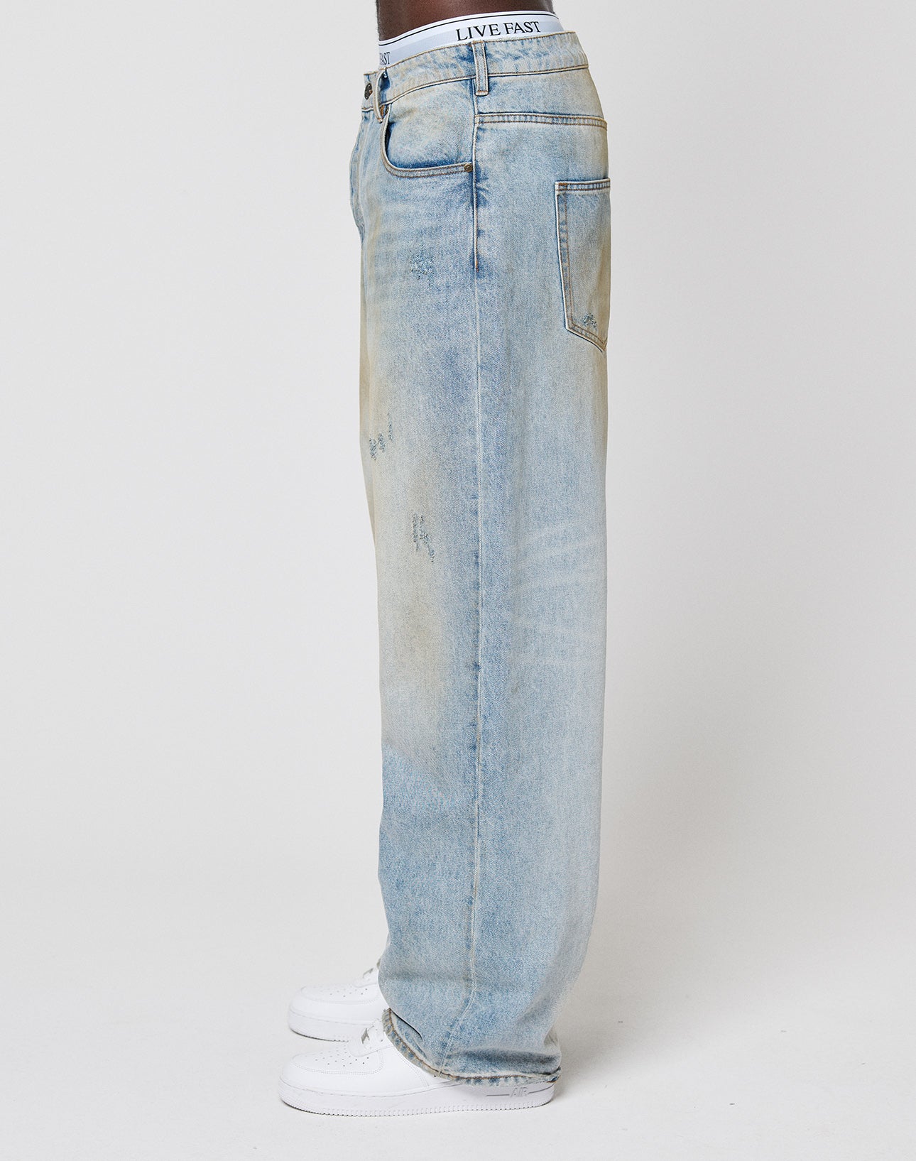 A person stands sideways in LFDY Baggy Denim by LIVE FAST DIE YOUNG—light-wash blue jeans with subtle distressing and a LIVE FAST waistband—paired with white sneakers against a plain white background.
