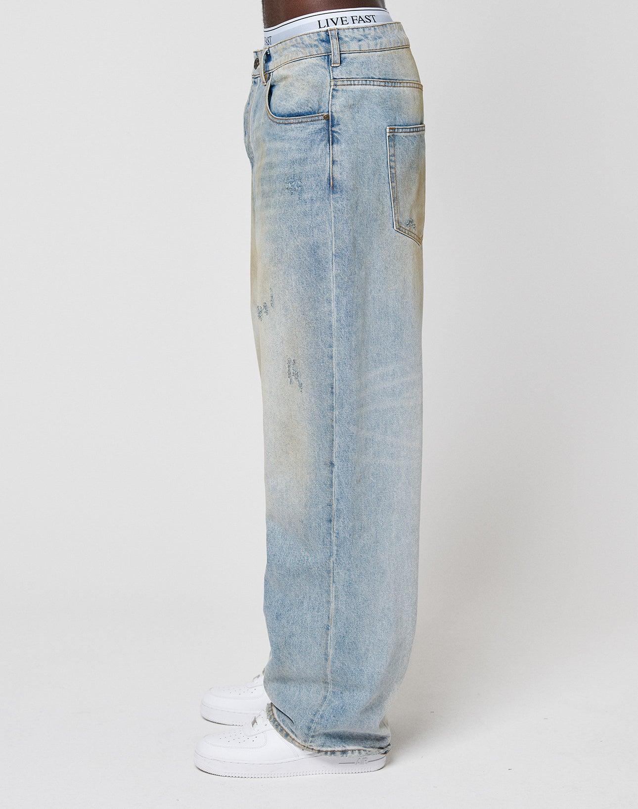 A person stands sideways in LFDY Baggy Dirt Denim by LIVE FAST DIE YOUNG—baggy, light blue, loose-fit jeans with a faded wash and straight leg—paired with white sneakers. The waistband label says LIVE FAST. The background is plain white.