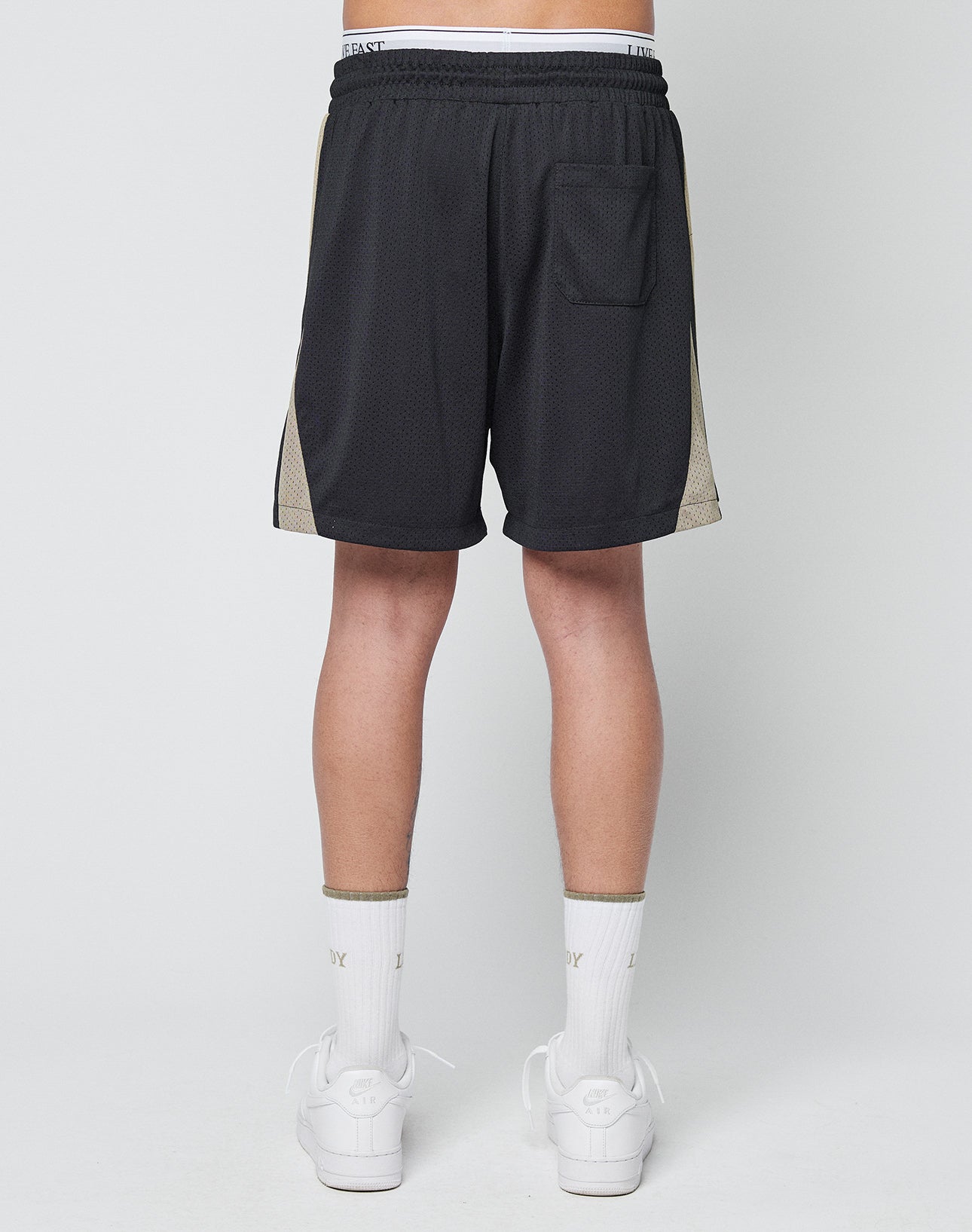 A person is facing away, wearing LIVE FAST DIE YOUNG Striped Mesh Shorts with a back pocket, paired with white socks and sneakers, against a plain light-colored background.