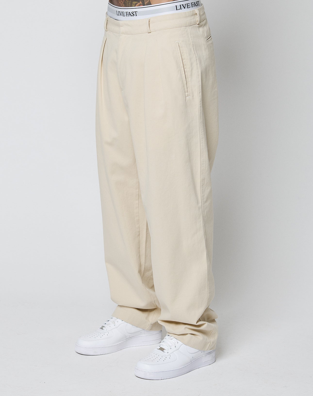 Model in beige Wide Chino trousers (100% Baumwolle, Doppel-Bundfalte) by LIVE FAST DIE YOUNG, with visible white LIVE FAST waistband underwear and white sneakers, standing against a plain white background. | model