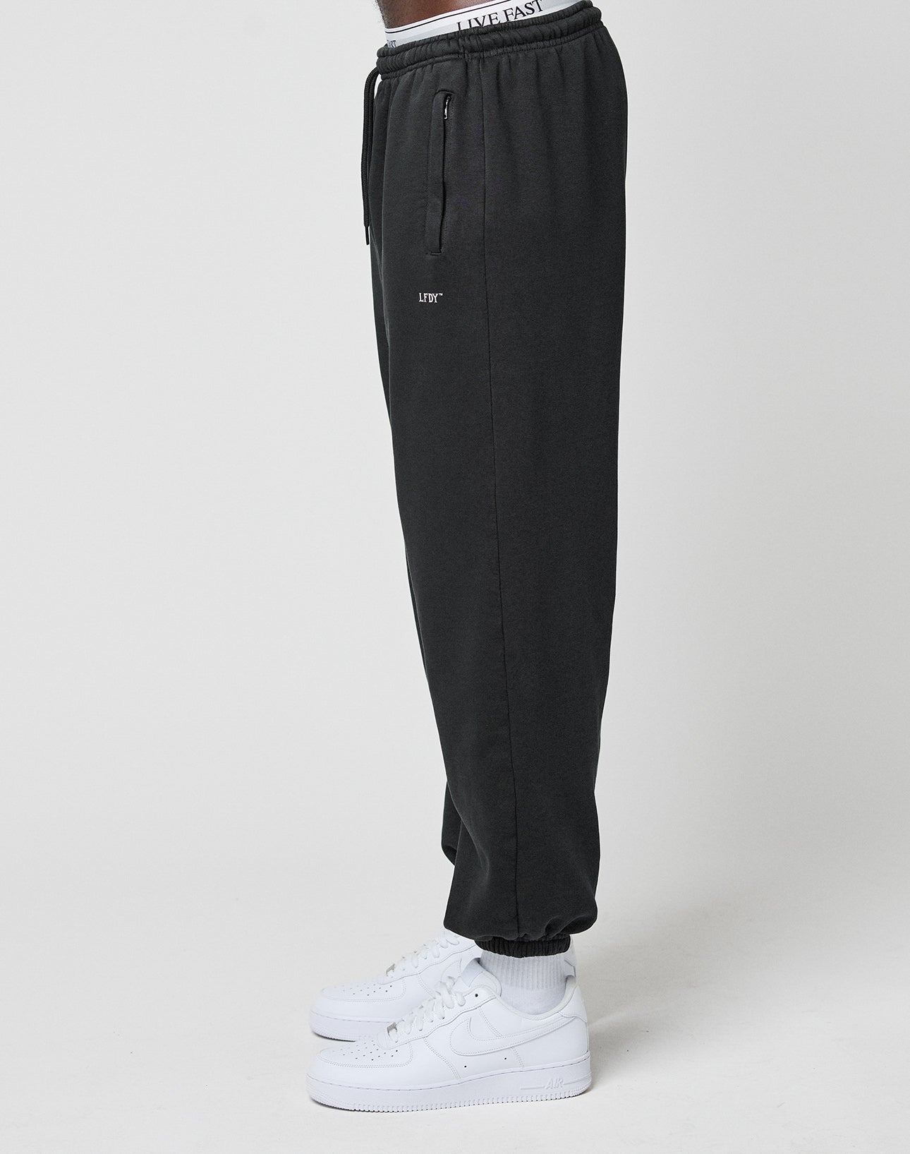A person in LIVE FAST DIE YOUNG Basic Loose Jogger sweatpants with zippered pockets, white undershorts showing a LIVE FAST waistband, and white Nike Air Force 1s, standing sideways against a plain white background.