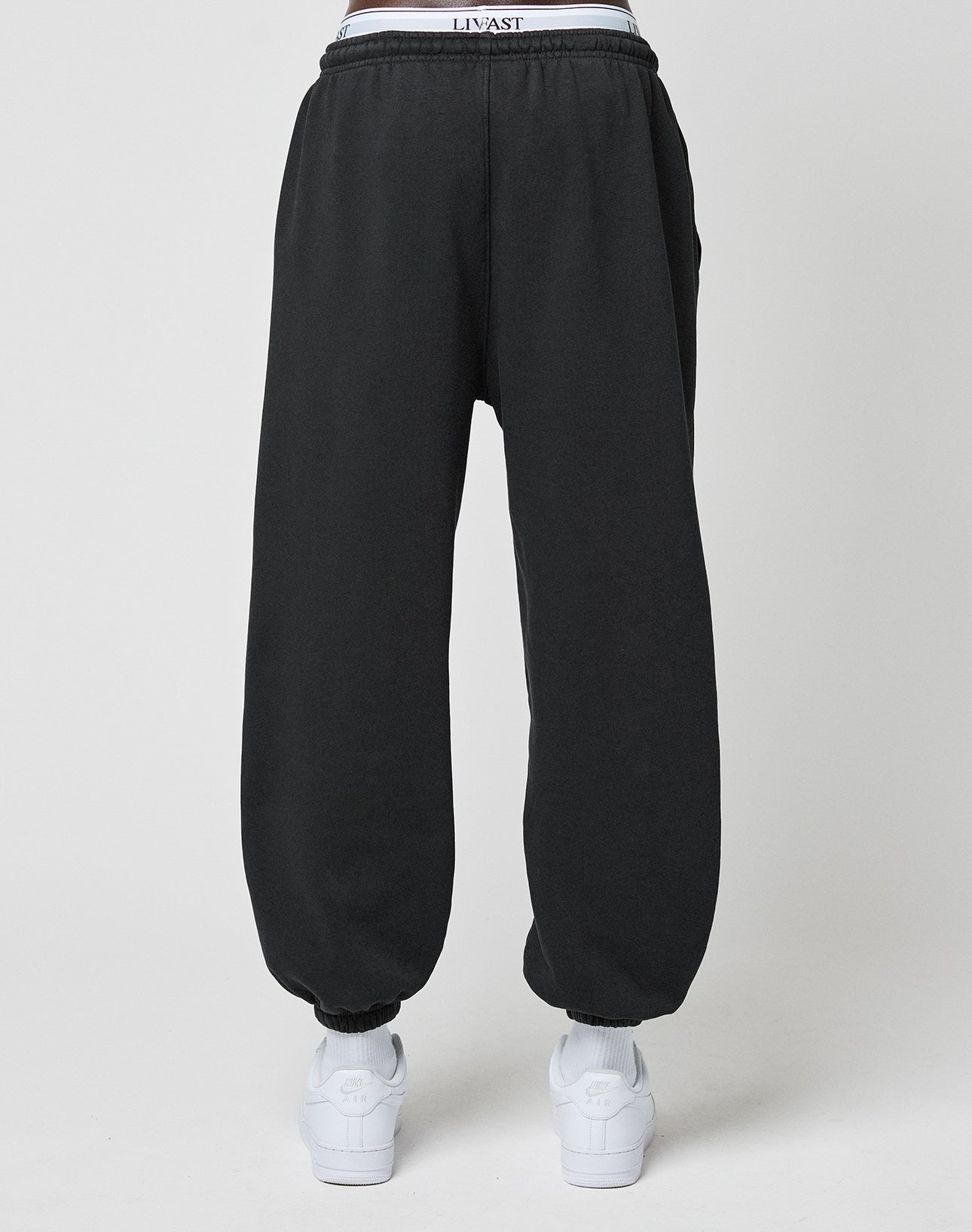 A person stands with their back to a light background wearing LIVE FAST DIE YOUNG Basic Loose Jogger sweatpants with an Elastik-Bund cuff, white sneakers, and a visible white LIVIAST logo print waistband.