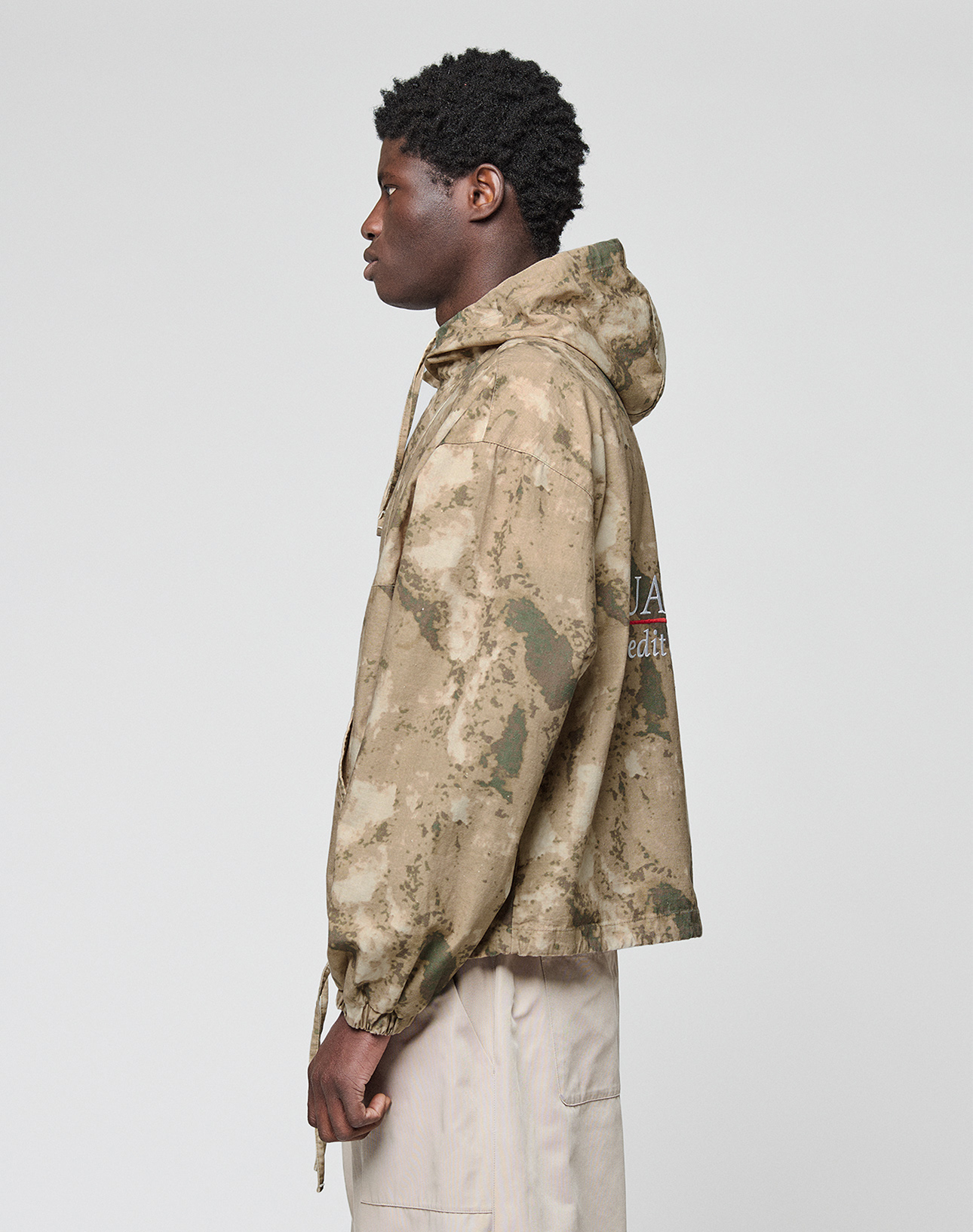 A person in profile faces left, wearing the LIVE FAST DIE YOUNG LFDY Camo Halfzip Jacket with an Oversize Fit and drawstrings, paired with pale pants, set against a plain light background.