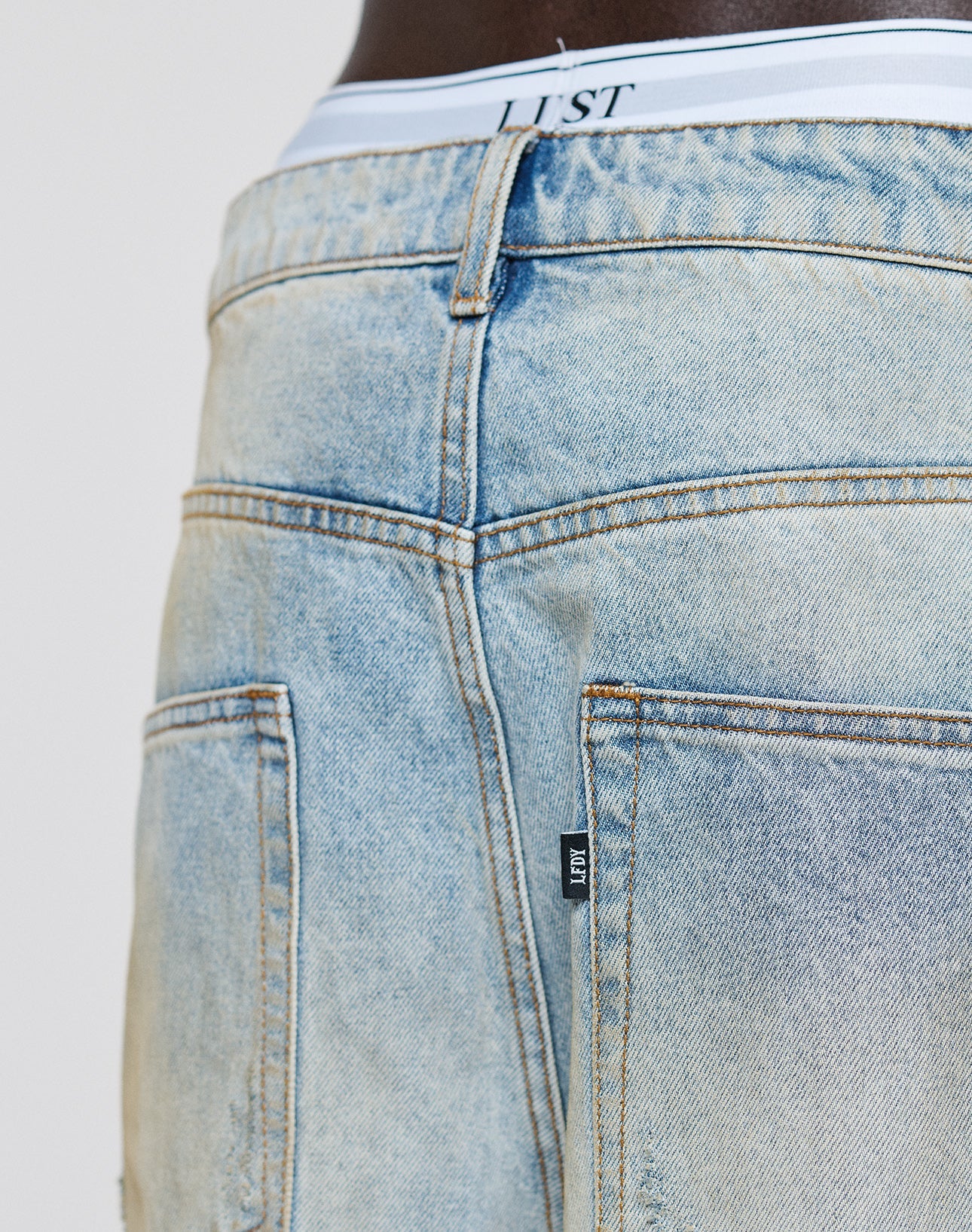 Close-up of the back of a person wearing LIVE FAST DIE YOUNG’s LFDY Baggy Denim in light blue, faded wash, with two back pockets and a white waistband marked LJST.