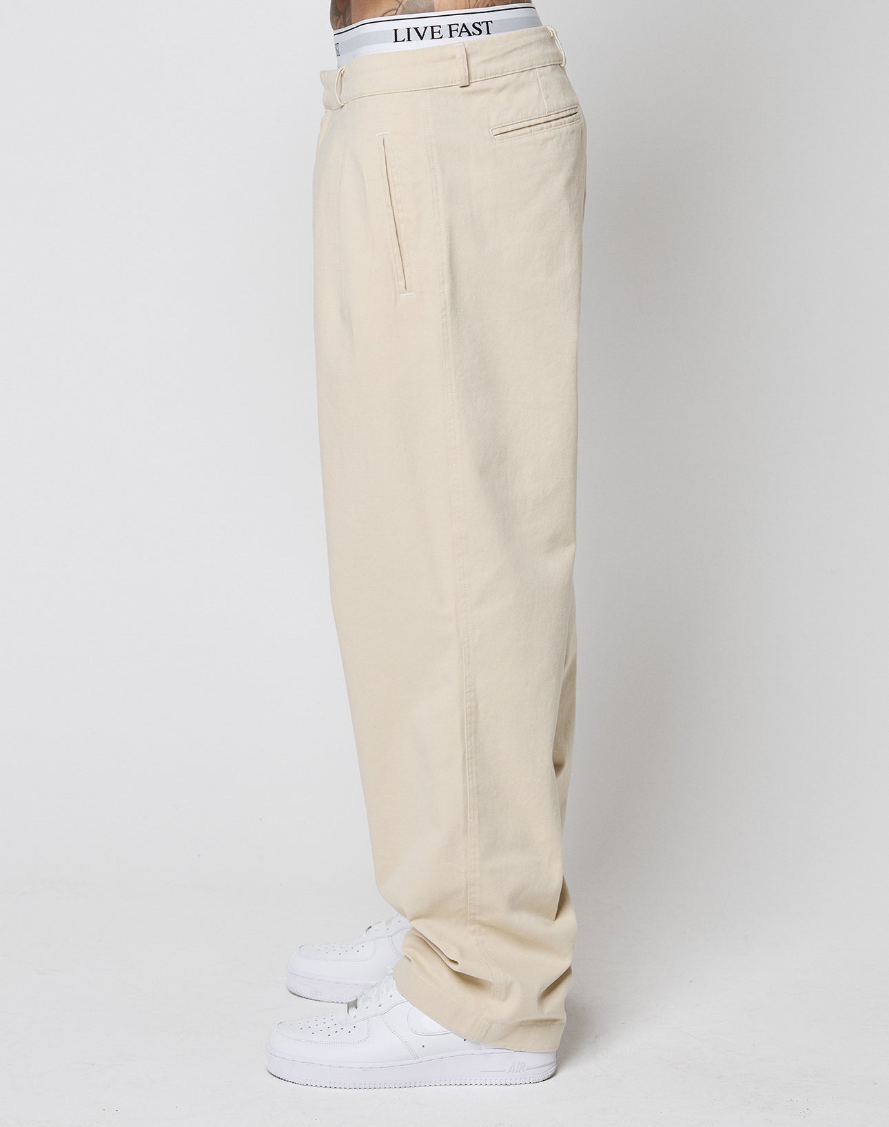 A person stands sideways in beige Wide Chino pants by LIVE FAST DIE YOUNG, styled with white sneakers and a white LIVE FAST waistband, against a plain white background.