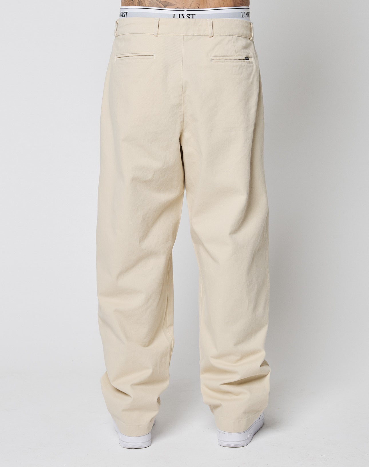 A person faces away, wearing LIVE FAST DIE YOUNGs Wide Chino—loose, off-white trousers with Doppel-Bundfalte, back welt pockets, a white waistband with text, and white sneakers. Made from 100% Baumwolle. Plain light background.