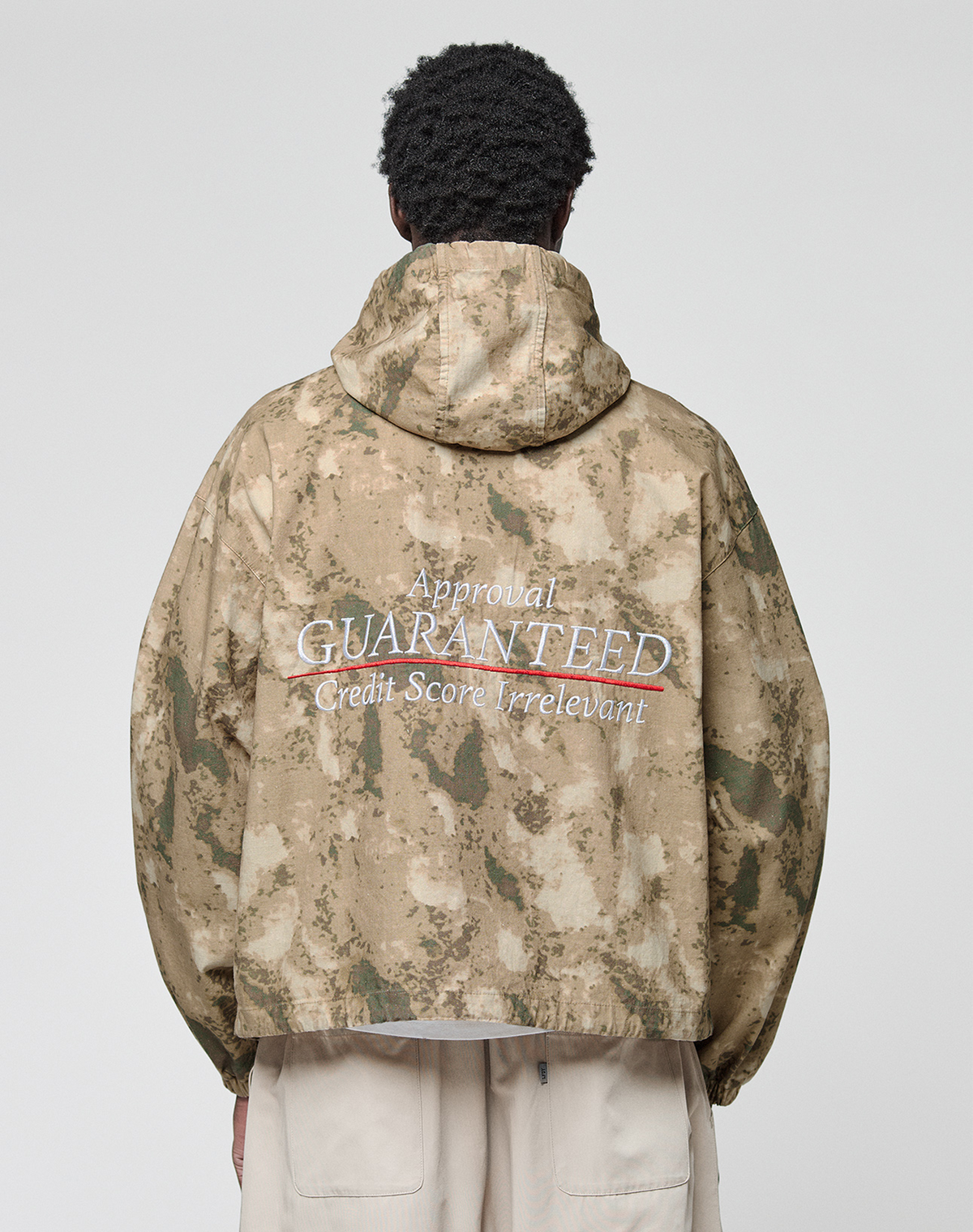 A person is seen from behind wearing the LIVE FAST DIE YOUNG LFDY Camo Halfzip Jacket, an oversize tan and green camouflage piece with a hood and “Approval GUARANTEED Credit Score Irrelevant” on the back, paired with light-colored pants.