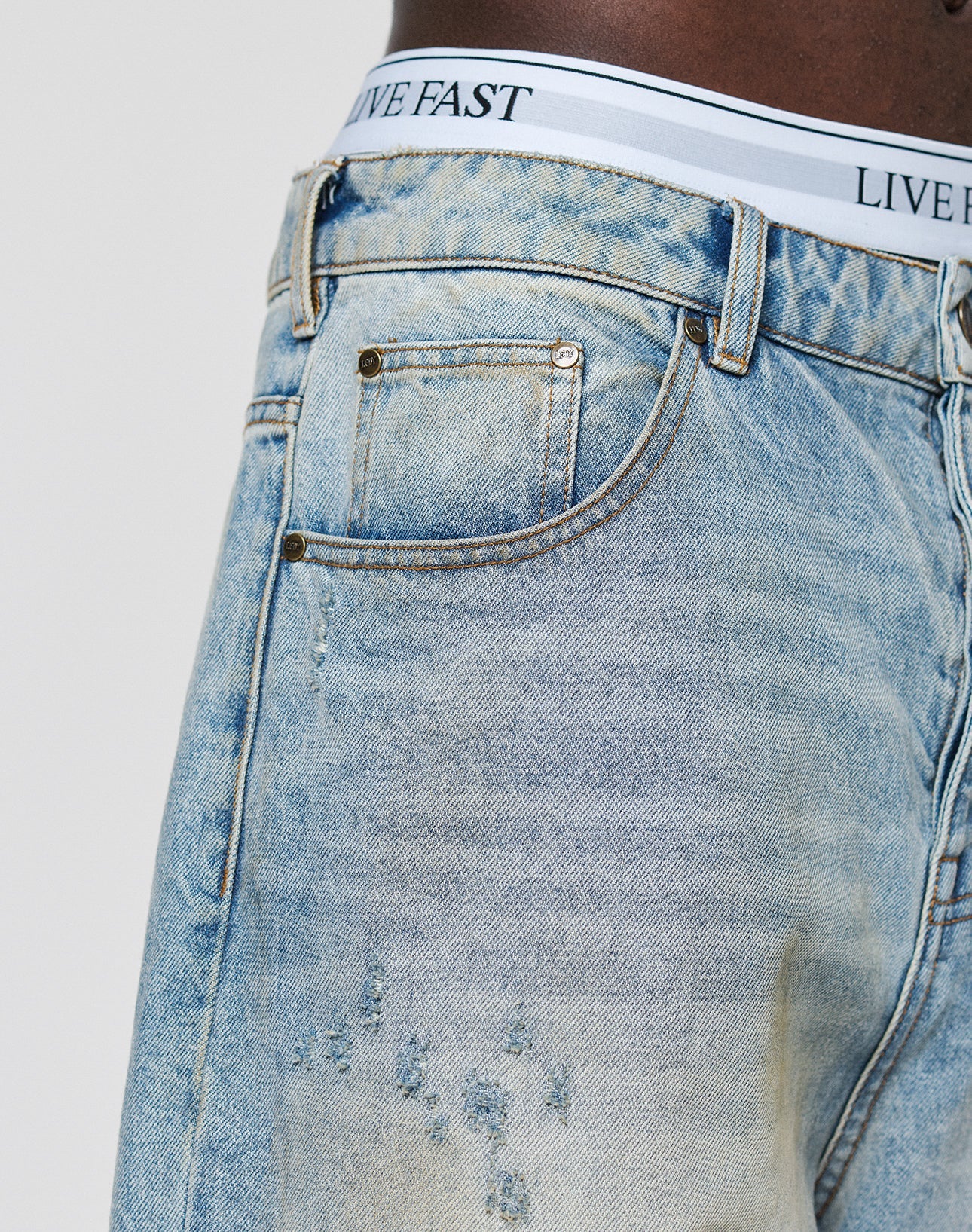Close-up of a person in LFDY Baggy Dirt Denim jeans by LIVE FAST DIE YOUNG, light blue and slightly distressed with a straight leg. Made from 100% Baumwolle, paired with white underwear featuring a LIVE FAST waistband.