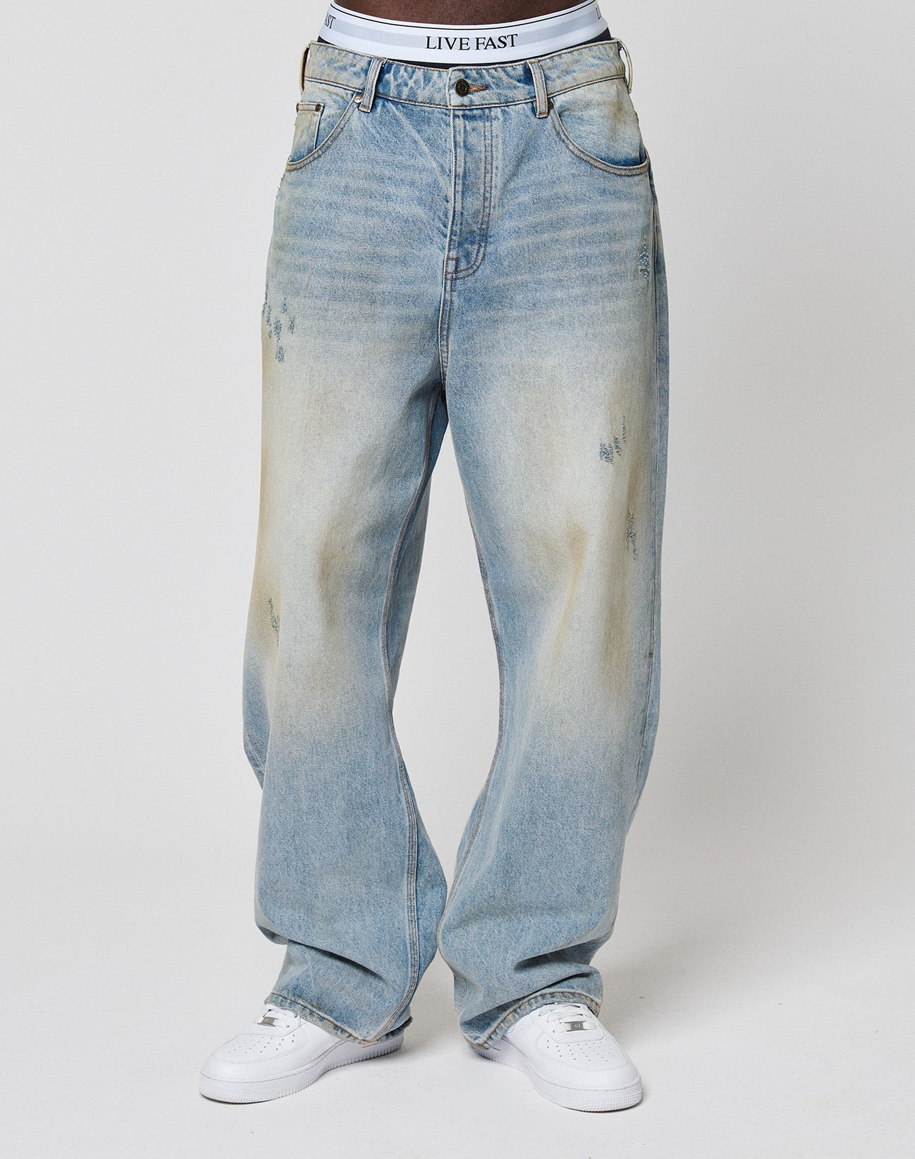 Someone wearing LFDY Baggy Dirt Denim jeans by LIVE FAST DIE YOUNG, styled baggy and faded blue with straight legs, white sneakers, and visible LIVE FAST branded white underwear, stands against a plain background.