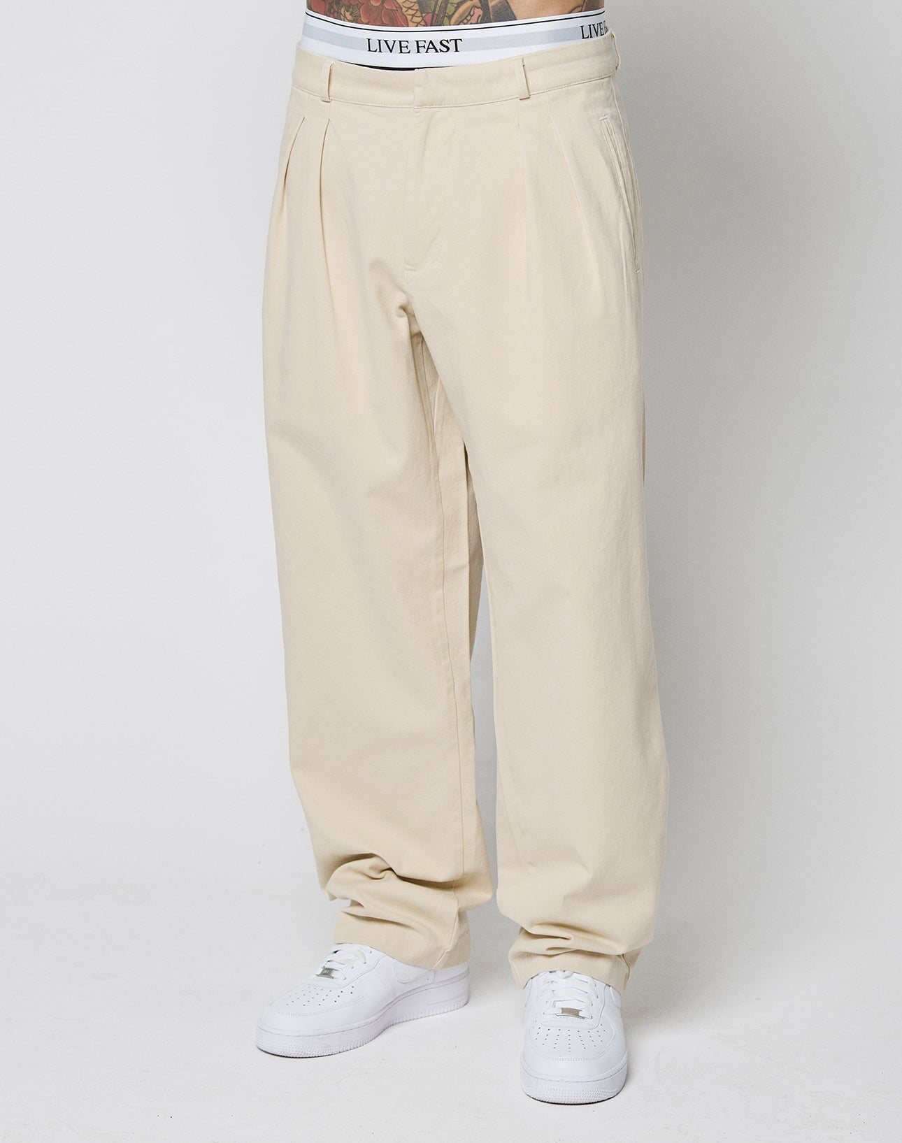 A person wears beige Wide Chino pants by LIVE FAST DIE YOUNG with Eingrifftaschen and pleats, paired with white sneakers and white underwear featuring a visible LIVE FAST waistband, standing against a plain white background.