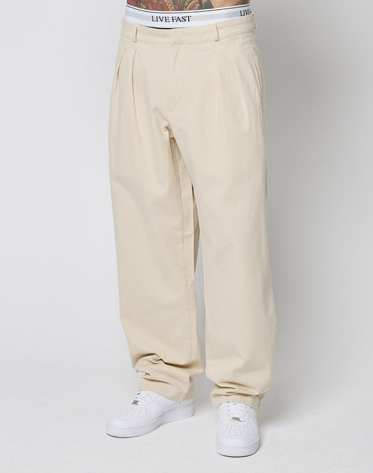 A person wears beige Wide Chino pants by LIVE FAST DIE YOUNG with Eingrifftaschen and pleats, paired with white sneakers and white underwear featuring a visible LIVE FAST waistband, standing against a plain white background.