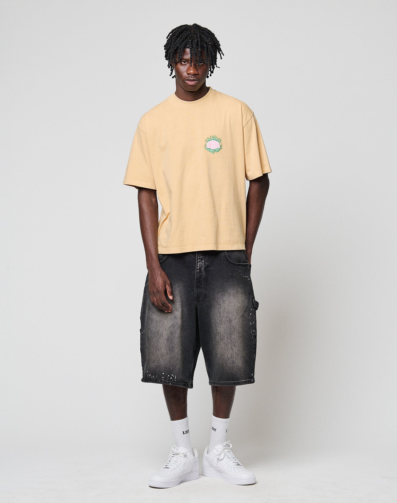 A young man wears the LFDY Flames Chest Tee by LIVE FAST DIE YOUNG—an oversized beige T-shirt with a small green and pink front print, paired with black baggy denim shorts, white crew socks, and white sneakers. Made from 100% Baumwolle.