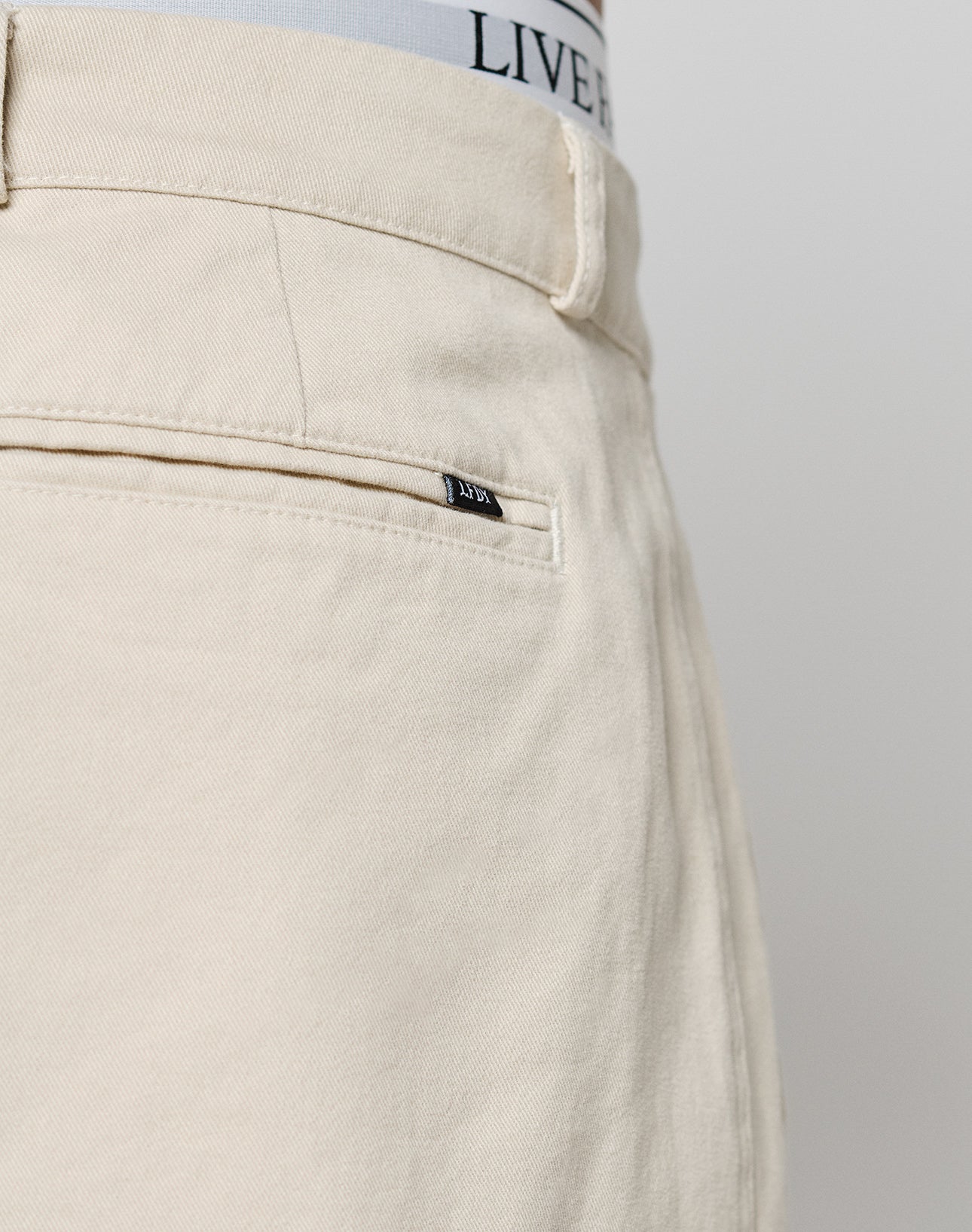 Close-up of the back of beige Wide Chino pants by LIVE FAST DIE YOUNG, in 100% cotton, showing a welt pocket with a black tag, part of a belt loop, and the waistband of LIVE FAST DIE YOUNG underwear visible at the top.