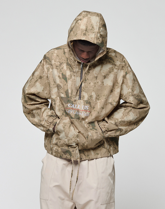 A person wears the LFDY Camo Halfzip Jacket by LIVE FAST DIE YOUNG, a tan and beige camo hooded jacket with "CALL US 49 Live Fast" on the front and an oversize fit. Hands in pockets, they face down against a plain white background.