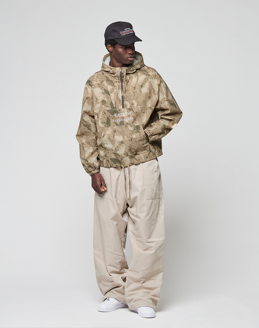 A person in a black cap, wide beige pants, white sneakers, and the LIVE FAST DIE YOUNG LFDY Camo Halfzip Jacket stands against a plain light gray background, looking to the side with one hand in their pocket.