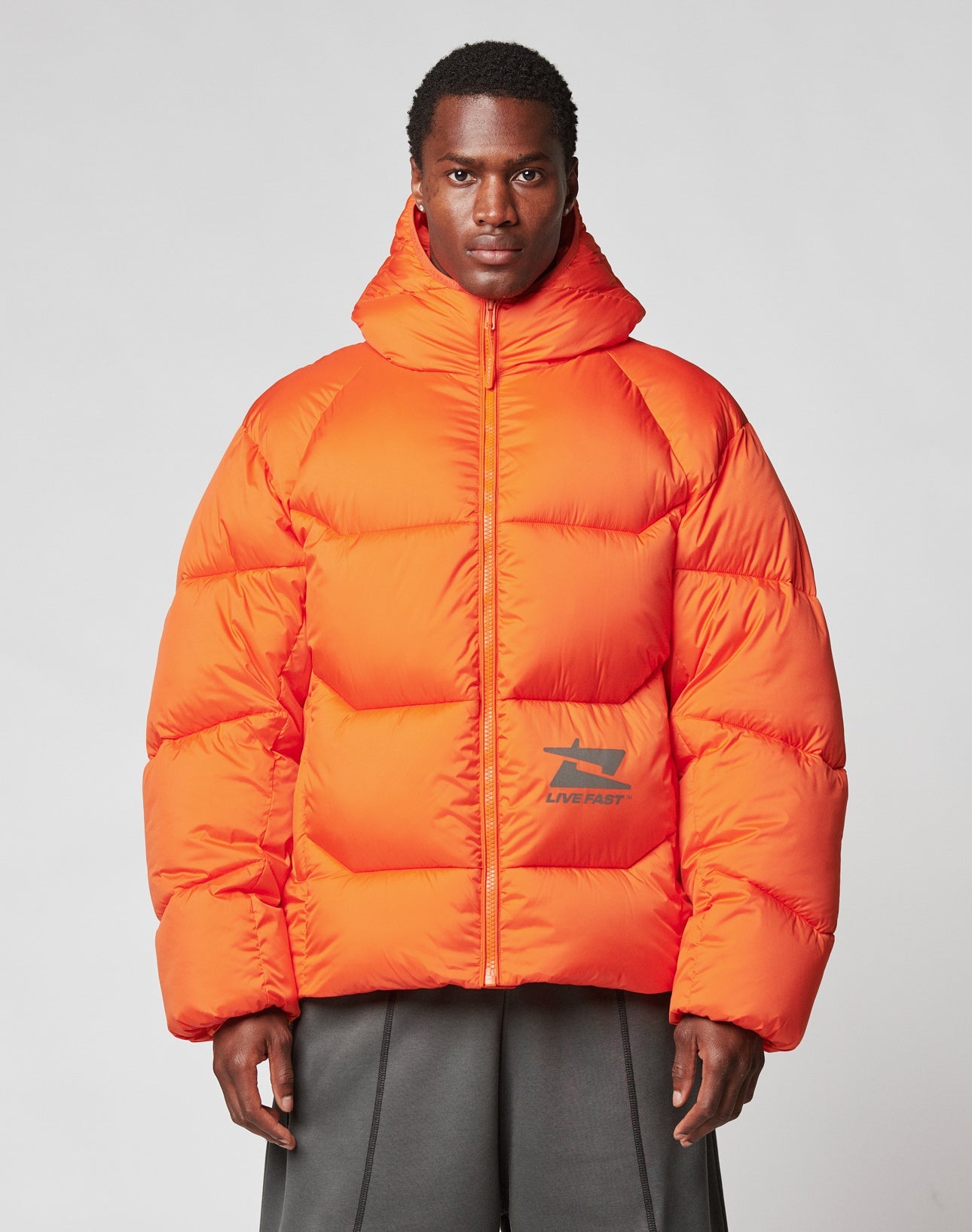 A person in the Live Fast Puffer Jacket by LIVE FAST DIE YOUNG, a bright orange oversized hooded style with a small black and red front logo, stands against a neutral background with a serious expression.