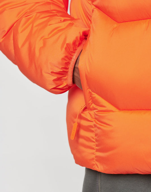 A close-up shows a hand in the pocket of the LIVE FAST DIE YOUNG Live Fast Puffer Jacket in bright orange, featuring visible stitching, an oversize fit, and a zipper pull against a plain light background.