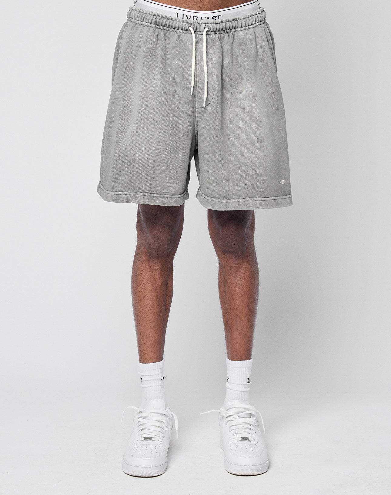 A person stands against a plain background wearing LIVE FAST DIE YOUNG Basic Sweat Shorts in grey with a regular fit, paired with white socks and sneakers. Their upper body is not visible in the image.